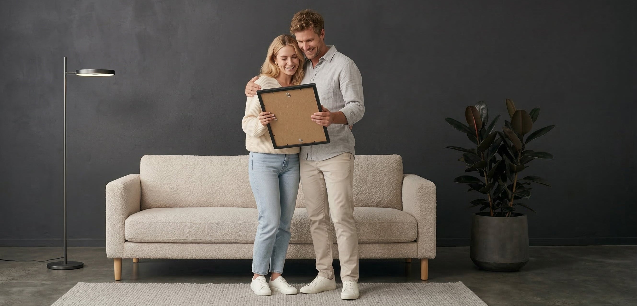 Man and woman holding a framed picture in a room with a beige sofa and dark walls.